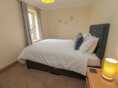 A bedroom with a bed white bedding a bedside table with lamp and a window with curtains at Whinny Cottage in Embleton