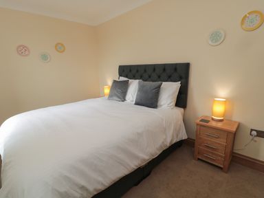 A bedroom with a double bed with white bedding and gray pillows a wooden nightstand with a lamp and decorative plates on the wall at Whinny Cottage in Embleton