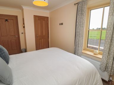 A bedroom with white bedding two wooden doors a window with patterned curtains and a small decorative item on the windowsill at Whinny Cottage in Embleton
