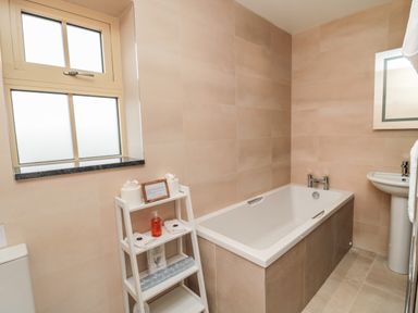 A bathroom with a bathtub a pedestal sink a small shelving unit with toiletries and a frosted window at Whinny Cottage in Embleton