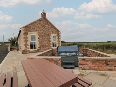 An outdoor patio with a hot tub in a sunken brick enclosure and a picnic table next to a brick house at Whinny Cottage in Embleton