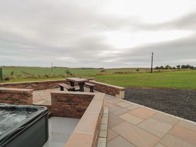 An outdoor patio area with a hot tub and a picnic table surrounded by a low brick wall overlooking fields at Whinny Cottage in Embleton