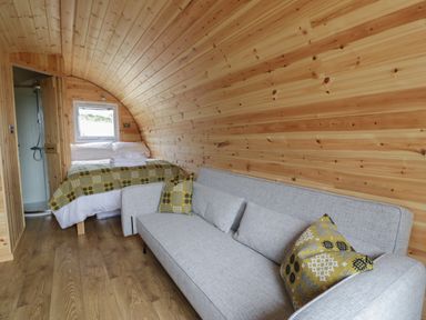 A curved wooden pod interior with a bed, grey sofa, patterned cushions and a bathroom at Dysynni Pod in Llanegryn near Tywyn