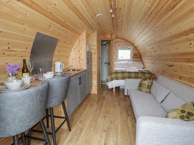 An interior view of a wooden pod with a kitchen area including a sink and kettle two grey bar stools a bed with blankets and pillows a grey sofa with cushions and a small window at Dysynni Pod in Llanegryn near Tywyn