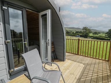A wooden deck with a chair and open doors leading to a pod with a field and hills in the background at Dysynni Pod in Llanegryn near Tywyn