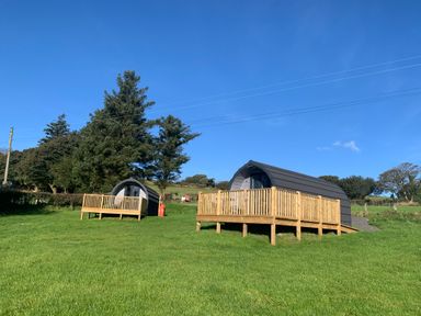 Two small black curved pods with wooden decks on a grassy field with trees in the background at Dysynni Pod in Llanegryn near Tywyn