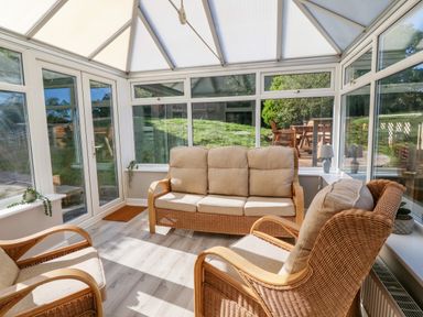 A conservatory with wicker chairs and a sofa with beige cushions and glass walls at Treestumps in Benllech