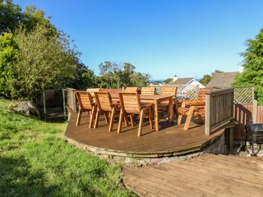 An outdoor wooden deck with a table and eight chairs surrounded by grass and trees at Treestumps in Benllech