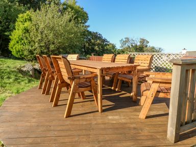 An outdoor wooden dining table with eight wooden chairs on a wooden deck with greenery and trees around at Treestumps in Benllech