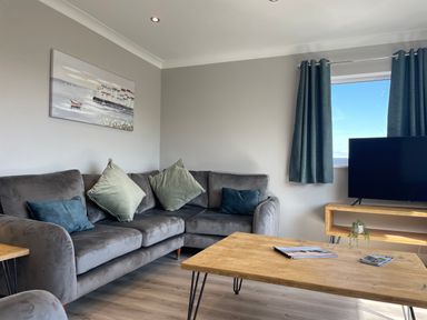 A living room with a grey sofa and blue green cushions a wooden coffee table with magazines a TV on a wooden stand and curtains at Treestumps in Benllech