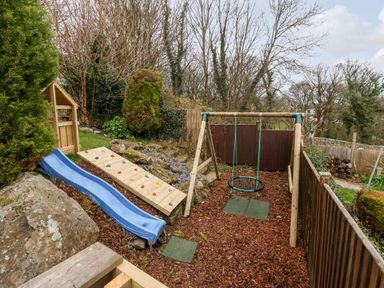 A backyard playground with a blue slide climbing wall and swing set surrounded by trees and wooden fences at Treestumps in Benllech