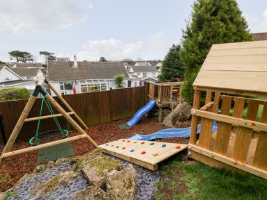 A backyard playground with a slide swing climbing wall and wooden playhouse at Treestumps in Benllech