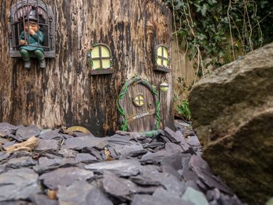 A miniature door and windows attached to a tree stump with a small figurine in a cage on the left and rocks in the foreground at Treestumps in Benllech