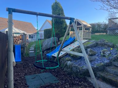A backyard playground with swings slides a climbing wall and a wooden playhouse at Treestumps in Benllech