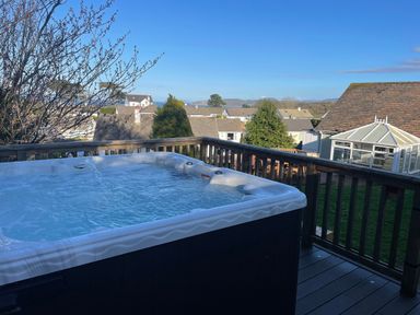 An outdoor hot tub on a wooden deck overlooking houses trees and a glass conservatory at Treestumps in Benllech