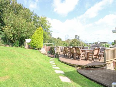 A garden with a wooden deck and table with chairs a small playhouse and a swing set at Treestumps in Benllech