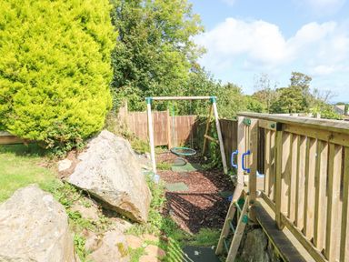 A wooden play structure with a swing and climbing ladder in a garden with rocks and trees at Treestumps in Benllech