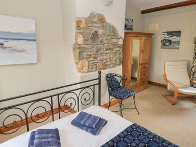 A bedroom with an iron bed frame, folded towels on the bed, a stone accent wall, a wooden wardrobe with mirror, a cushioned chair, and a lounge chair at 2 Yr Hen Felin in Castle Morris near Letterston