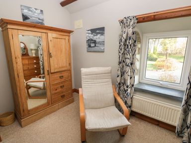 A room with a wooden wardrobe with a mirror a chair with light cushioned seat and back and a window with patterned curtains at 2 Yr Hen Felin Castle Morris near Letterston