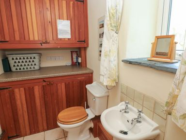 A bathroom with a toilet and sink next to wooden cabinets and a window with floral curtains at 2 Yr Hen Felin Castle Morris near Letterston