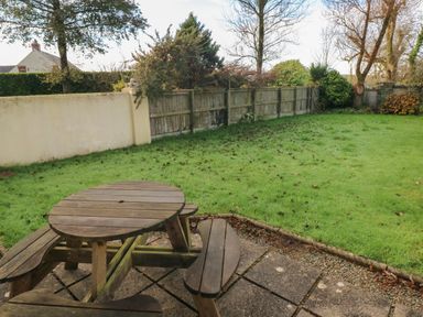 A backyard with a wooden picnic table on a stone patio fenced by a wooden and concrete wall at 2 Yr Hen Felin in Castle Morris near Letterston