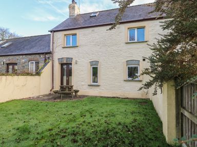 A backyard with grass and a wooden picnic table next to a cream stone house at 2 Yr Hen Felin in Castle Morris near Letterston