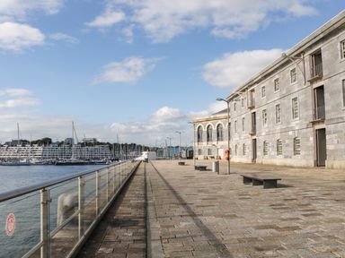 A waterfront promenade with stone paving benches and a stone building next to a marina with boats and yachts at Clarence Riverside Royal William Yard in Plymouth