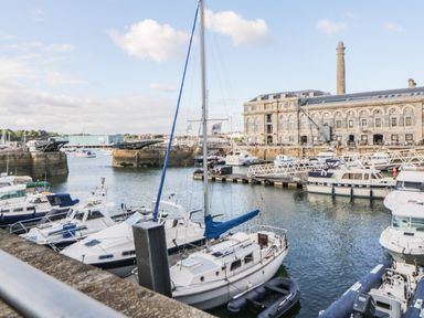 A marina with multiple boats docked and a large stone building with a tall chimney in the background at Clarence Riverside Royal William Yard in Plymouth