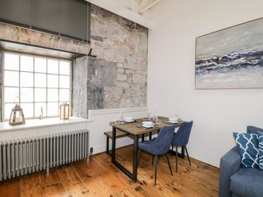 A dining area with a wooden table set for four with blue chairs beside a large window and exposed stone wall at Clarence Riverside Royal William Yard in Plymouth