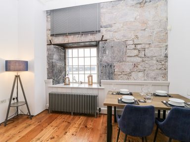 A dining area with a wooden table set for four and blue chairs near a window with stone walls and a floor lamp at Clarence Riverside Royal William Yard in Plymouth