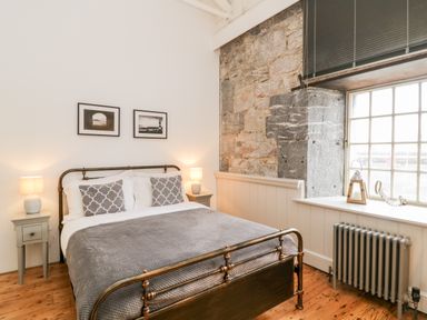 A bedroom with a metal bed frame two bedside tables with lamps and a large window with a stone wall at Clarence Riverside Royal William Yard in Plymouth