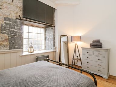 A bedroom with a grey bedspread a stone window frame a standing mirror a floor lamp and a chest of drawers at Clarence Riverside Royal William Yard in Plymouth