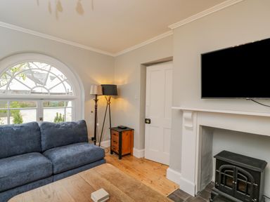 A living room with a blue sofa a wooden coffee table a black stove a television on the wall and a window at White Roses Whitchurch near Monmouth