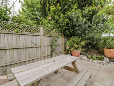 A wooden picnic table on a paved patio with a wooden fence and potted plants in the garden at White Roses Whitchurch near Monmouth