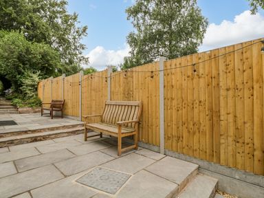 An outdoor paved patio area with two wooden benches and a wooden fence with string lights at White Roses in Whitchurch near Monmouth