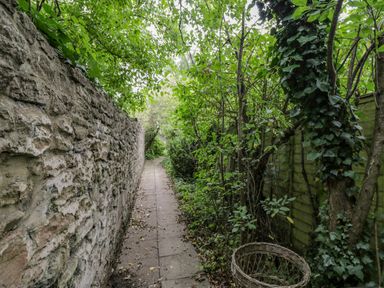 A narrow paved path between a stone wall and wooden fence with green trees and bushes at White Roses in Whitchurch near Monmouth