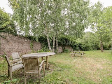 A garden with wooden chairs and a table under a tree by a stone wall at White Roses in Whitchurch near Monmouth