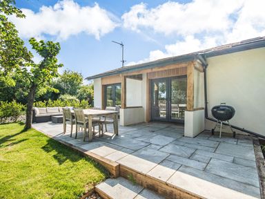 A patio with a wooden dining table and chairs a barbecue and an outdoor sofa next to a garden at The Paddock in Honington near Shipston-On-Stour