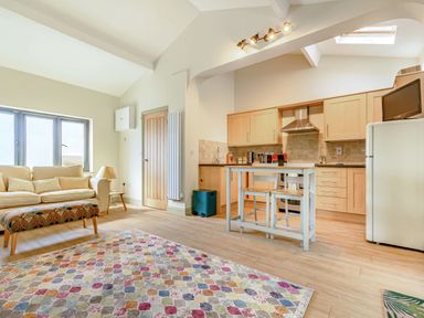 An open-plan living room and kitchen with a sofa, colorful rug, small table with two chairs, wooden cabinets, and a refrigerator at The Paddock in Honington near Shipston-On-Stour