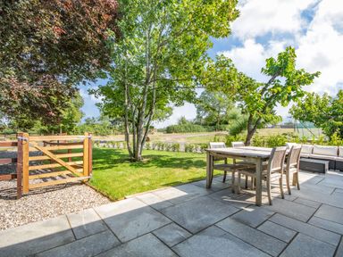 An outdoor patio area with a wooden dining table and chairs next to a grassy lawn with trees at The Paddock in Honington near Shipston-On-Stour