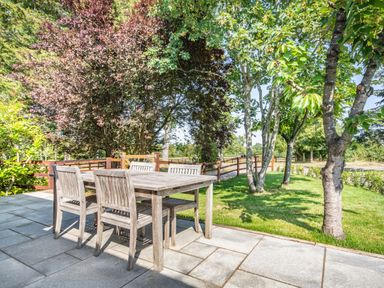 An outdoor patio with a wooden table and chairs surrounded by trees and grass at The Paddock in Honington near Shipston-On-Stour