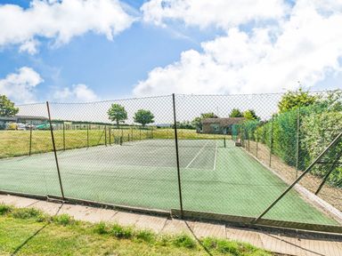 An outdoor tennis court enclosed by a chain link fence surrounded by grass and trees at The Paddock in Honington near Shipston-On-Stour