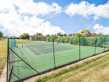 An outdoor tennis court surrounded by a green fence with houses and trees in the background at The Paddock in Honington near Shipston-On-Stour