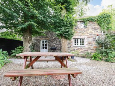 A stone house with a wooden picnic table and trees in an outdoor gravel area at The Cottage in St Kew