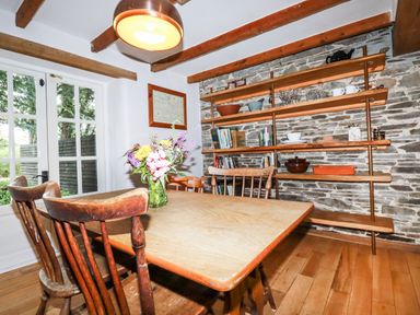 A dining room with wooden table and chairs a vase with flowers wooden shelves with books and dishes and a stone wall at The Cottage in St Kew