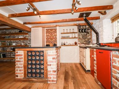 A kitchen with wooden beams brick island with wine rack shelves with glassware and red cabinets at The Cottage in St Kew