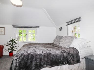 A bedroom with a bed covered in gray blankets and pillows next to two windows with blinds and a plant in a red pot at The Cottage in St Kew