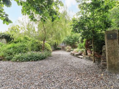 A gravel pathway surrounded by trees and plants leading to a building with a window at The Cottage in St Kew