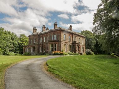 A large brick house with multiple chimneys on a curved driveway surrounded by grass and trees at The Residence in Cleator