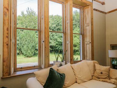 A living room with a beige sofa and cushions near three large wooden framed windows with plants on the windowsill at The Residence in Cleator